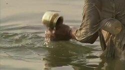 A man repeatedly dips a small brass pot into a river in India. Stock Footage
