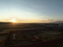Yellow sun setting behind hills with blue skies,  with buildings, walls and fields in f/g, Tiwanaku Tiahuanaco/Tiahuanacu, Bolivia Stock Footage