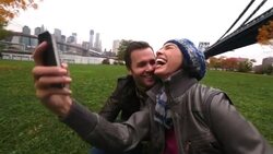 Young woman takes smartphone photo with boyfriend as he kisses her cheek; New York skyline in background Stock Footage