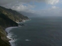 Accelerated timelapse. A wide frame shows waves crashing against high cliffs in Big Sur. White clouds drift across a blue sky, cast moving shadows against the cliffs. Stock Footage