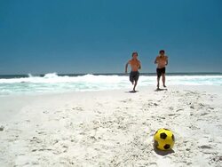 Slow motion wide shot two men running on beach and kicking soccer ball Stock Footage