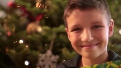 Young boy with present looks up and smiles at camera in front of Christmas tree Stock Footage