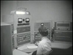 A woman works in a radio studio control room as F.B.I. agents watch from behind a glass. News Clip