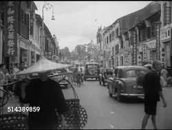 1953: KUALA LUMPUR, MALAYA: CHINATOWN: WS Street scene w/ Asian male in large conical straw hat carrying baskets on shoulder pole, cars moving up street lined w/ shops. Angled WS Sultan Abdul Samad Building w/ Medan Merdeka (Merdeka Square) fountain FG. Instructional Video