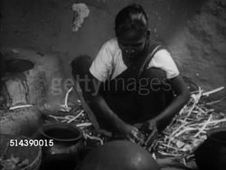 1949: INDIA CASTE SYSTEM: OUTCASTS / UNTOUCHABLES: VS Mother preparing meal, MS Soaking, turning rice in bowl with hands, eldest son Ezdoss watching, mother stirring cooking pot over fire inside one room hut.    Indian, indigenous, poverty. Instructional Video