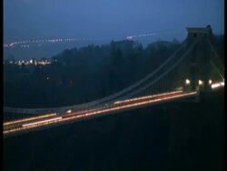 T/L Bristol Clifton Suspension Bridge at Night - Pan left across Bridge, streaky traffic, city lights in background, bridge lights come on Stock Footage