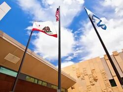 LA T/L flag poles with USA and California flags flapping in the wind against blue sky and modern public library building /Rancho Mirage, California, USA Stock Footage
