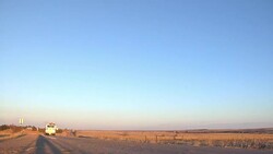 Semi truck carries a backhoe on a trailer and drives down a rural highway and passes a farm tractor discing a field as the sun sets. Stock Footage