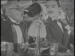 Two men in tuxedos talk at a table while Josephine Baker dances on stage in a bikini top and banana skirt. News Clip