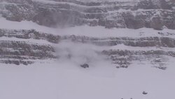 Snow falls over a sheer cliff toward a stand of evergreens. Stock Footage