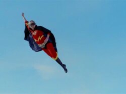 Wide shot young blonde woman in mask, cape, and superhero costume in midair simulating flying backwards after jumping on trampoline Stock Footage