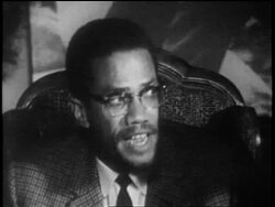 B/W 1962 close up Malcolm X with eyeglasses sitting in chair + speaking about Africa / documentary Stock Footage