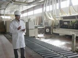 WS TS man in lab coat and hard hat walking along furniture production line, taking notes on handheld electronic device Stock Footage