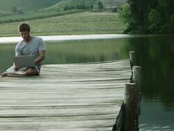 WS DS of man sitting on jetty by lake using laptop Stock Footage