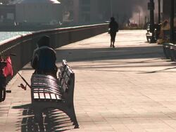 Man smoking on a park bench while fishing in the east river as a jogger runs past with steam rising in the background in manhattan at mid day Stock Footage