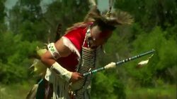 Native American Indian male wearing ceremonial clothes, feathered headdress and carrying Talking Stick performing war dance Stock Footage