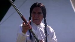Older seated male Native American Indian cleaning and pointing rifle with smoke in foreground and teepee behind Stock Footage