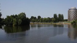 Leafy trees shade a quiet pond near a modern, cylinder-shaped office building. Stock Footage