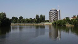 A cylinder-shaped steel building overlooks a quiet pond lined with leafy trees. Stock Footage