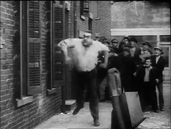 B/W 1917 large man (Eric Campbell) chasing policeman Charlie Chaplin around while crowd watches Stock Footage