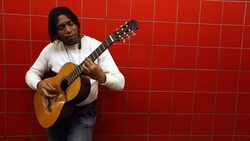 Street artist leans against red tile wall and plays acoustic guitar In urban subway station Stock Footage