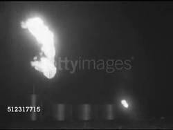 1952: DRILLING SAFETY: NIGHT: WS Flare stack (guyed supported) flame burning, fire, burning off excess gas or oil at refinery w/ tanks below, smaller flame on side, wind blowing smoke into lens, person walking by tank FG.    Rig, refinery, industrial Instructional Video