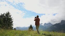 Middle aged man walks through mountain meadow and takes a picture with his tablet Stock Footage