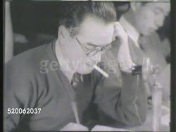 1942: JAPAN: NEWSPAPER: WS Newspaper building, pedestrians & automobiles on street outside. INT VS Journalists working at tables, reading papers, one male w/ cigarette in mouth writing, checking map, poster on wall, men working. Journalism, propaganda Instructional Video