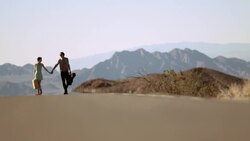 Young couple hold hands walking up a sweltering desert road Stock Footage