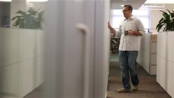 Office worker with coffee mug walks down hall, stops in doorway to chat (dolly shot) Stock Footage