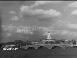 1951: HARVARD UNIVERSITY CAMPUS: XWS Harvard University campus Dunster House Tower (& clock) w/ Weeks Footbridge & Charles River FG. Male crew rowing on river w/ campus BG. Two male students walking, talking on campus sidewalk. Iconic, Ivy League. Instructional Video