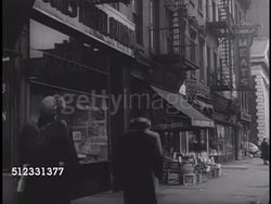 1950: TRAVEL AGENT STORE: Woman standing in front of travel agent storefront near fruit & meat market, people walking sidewalk. VS Travel Agency window display posters, brochures, '1950 Holy Year' advertising in window, 'Fly to Rome B O A C' Instructional Video