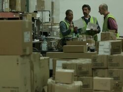 Three warehouse employees look at plans and talk while additional employee walks past in the foreground. Stock Footage