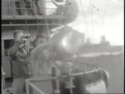 A sailor sends Morse code via a signal light while a second sailor stands behind him looking through binoculars. News Clip