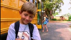 A young boy smiles as students get on and off of the school bus behind him. Stock Footage