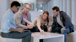 Creative young business people write and discuss ideas around white-board table in large corporate office common area (dolly shot) Stock Footage
