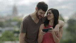 Young couple look at smartphone and laugh overlooking city skyline Stock Footage