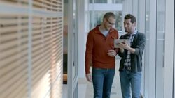 Male and female business partners write on white-board, two trendy businessmen discuss strategy with tablet as they walk up office hallway (dolly shot) Stock Footage