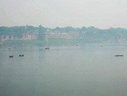 Rowing boats transport people in hazy misty distance on silvery river with big ancient fortifications as backdrop. Kumbh Mela, India Stock Footage