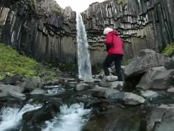MS shot of river below Svartifoss waterfalls, hanging hexagonal basalt columns underlying and young woman walking up to falls and taking photo / Skaftafell National Park, Austur-Skaftafellssysla, Iceland   Stock Footage