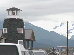 "Lighthouse of The Salty Dawg Saloon with light active, cars passing both directions, Homer Spit, Homer, Kenai Peninsula, Alaska, snow capped mountains of Kachemak Bay State Park and Wilderness Park visible." Stock Footage