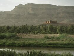 A river runs in the foreground, whilst thick Moroccan vegetation leads to an old building dwarfed by mountains. Stock Footage