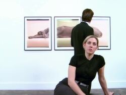 Dolly past woman sitting on bench at gallery opening and looking at artwork across room / man looking at three-panel print of a Weimaraner by William Wegman on wall behind woman Stock Footage