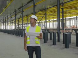 MS engineer holding plan standing in large hangar, looking around ostensible checking things, front view, RED R3D 4K Stock Footage