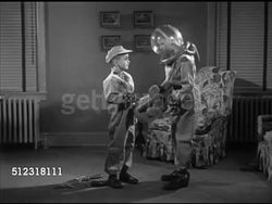 1953: DRAMATIZATION: PLAYING ASTRONAUT: WS Two boys standing in living room dressed in spacesuits, taller boy in round plastic helmet placing shoulder resting longer helmet on younger shorter boy. Home Movies, brothers, siblings, space, imagination. Instructional Video