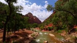 Camera tilts up from a fast running river to red-rock mountain tops against blue cloud-filled skies with a sun flare Stock Footage