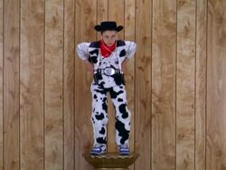 Medium shot boy in cowboy costume on pedestal aiming toy pistols w/wood paneling in background Stock Footage