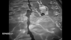 1950: COMMUNITY POOL: Woman in two-piece bathing suit passing 'Bathers Must Use Showers....' sign, Many people in, around pool, diving off diving board, swimmers swimming underwater, people playing. Lifeguard blowing whistle. Couple sitting on edge. Instructional Video