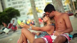 Young Brazilian couple pose for smartphone selfie on beach by crowded Rio street Stock Footage