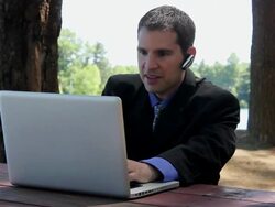 MS Man sitting on table and talking on bluetooth while typing on laptop near lake / Portland, ME, United States Stock Footage